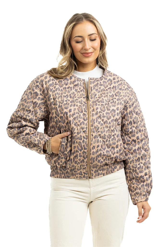 A woman smiles with her eyes closed, wearing the Katydid Brown Leopard Quilted Jacket and white pants, standing with one hand in her pocket against a plain white background.