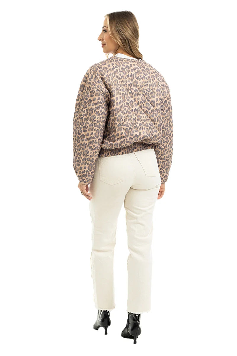 A woman with long light brown hair faces away, wearing a Katydid Brown Leopard Quilted Jacket, off-white jeans, and black ankle boots against a white background.