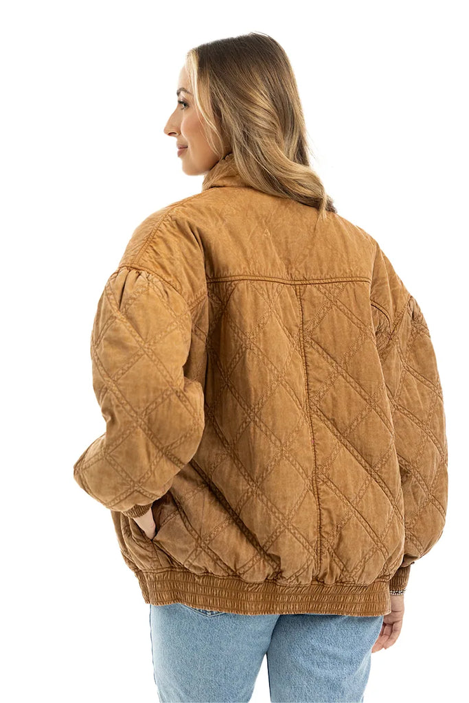 A woman with wavy blonde hair models the Katydid Brown Luxe Garment Washed Oversized Jacket, featuring a diamond quilted pattern, paired with blue jeans as she stands looking left against a white background.