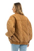 A woman with wavy blonde hair models the Katydid Brown Luxe Garment Washed Oversized Jacket, featuring a diamond quilted pattern, paired with blue jeans as she stands looking left against a white background.