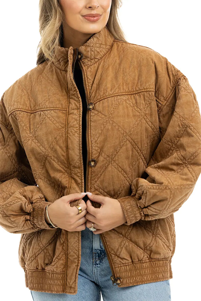 A woman wearing the Katydid Brown Luxe Garment Washed Oversized Jacket with a high collar and elastic cuffs, paired with light blue jeans. She smiles slightly and clasps the brown oversized jacket at the front.