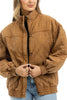 A woman wearing the Katydid Brown Luxe Garment Washed Oversized Jacket with a high collar and elastic cuffs, paired with light blue jeans. She smiles slightly and clasps the brown oversized jacket at the front.