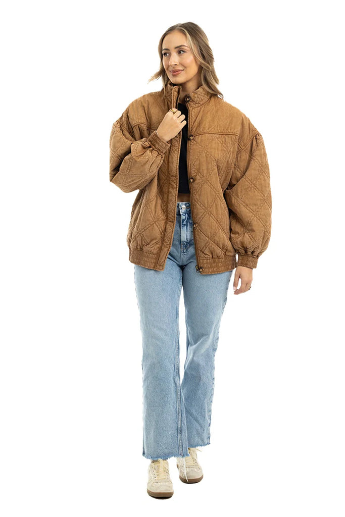 A woman models the Katydid Brown Luxe Garment Washed Oversized Jacket against a white background, pairing it with light blue jeans, a black top, and white sneakers. She has long wavy hair and smiles gently while looking to the side.