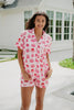 A woman smiles outdoors in front of a white house, wearing the Katydid Girly Cakes PJ Shorts Set decorated with pink and red cake illustrations.