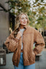 A woman with long blonde hair wears the Katydid Brown Luxe Garment Washed Oversized Jacket over a white top and blue jeans. She stands outdoors with greenery and buildings behind her, looking slightly left and smiling gently.