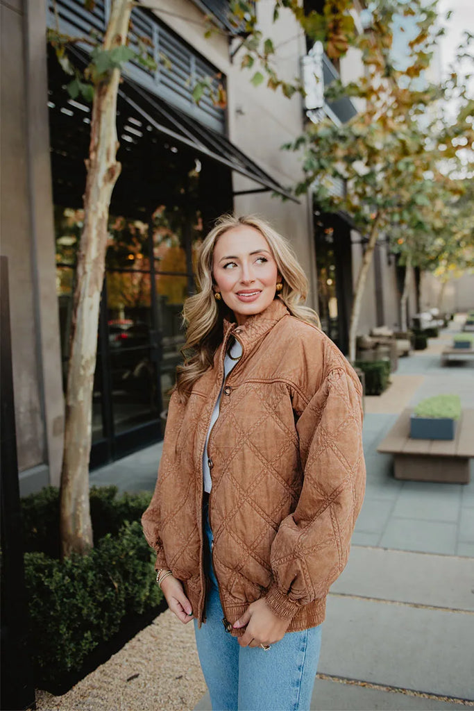 A woman with long blonde hair wears the Katydid Brown Luxe Garment Washed Oversized Jacket and blue jeans, standing outdoors on a city sidewalk with trees, benches, and modern buildings in the background.