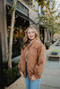 A woman with long blonde hair wears the Katydid Brown Luxe Garment Washed Oversized Jacket and blue jeans, standing outdoors on a city sidewalk with trees, benches, and modern buildings in the background.