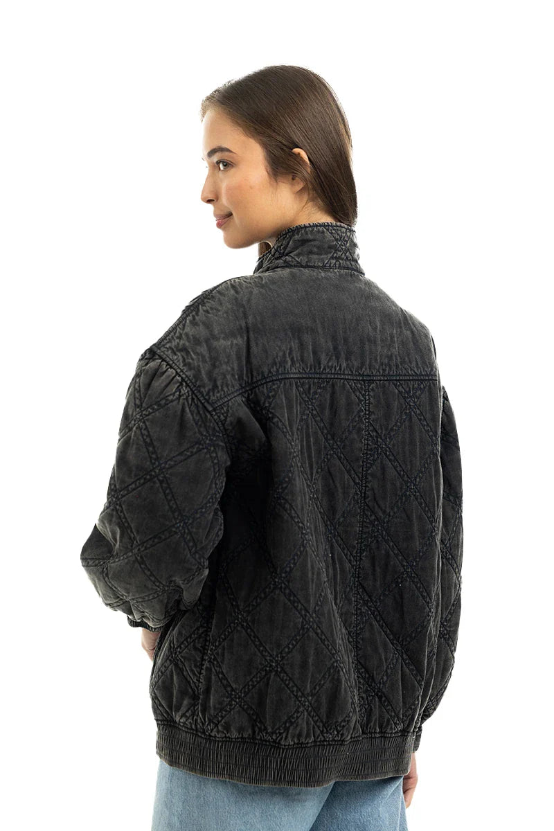 A woman with straight brown hair poses in the Katydid Womens Charcoal Luxe Oversized Jacket and light blue jeans, showcasing the jackets diamond quilting and relaxed vintage-inspired style against a white background.