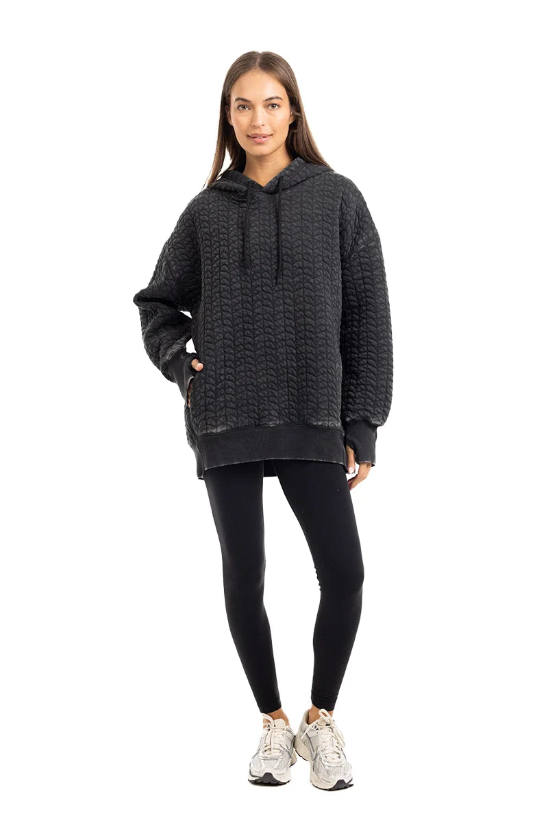 A woman with long straight hair stands against a white background, wearing the Katydid Charcoal Heart Quilted Pullover Hoodie, black leggings, and white sneakers. She looks relaxed, with one hand in her hoodie pocket.