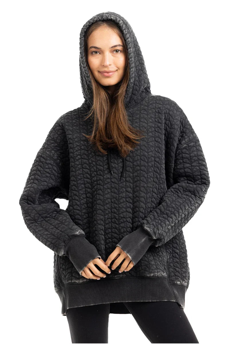 A woman with long brown hair wears the Katydid Charcoal Heart Quilted Pullover Hoodie and black leggings, smiling at the camera against a white background.