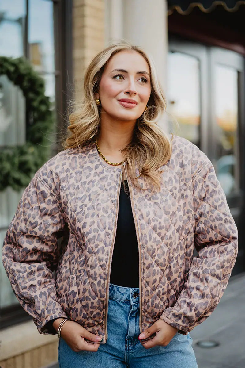 A woman with wavy blonde hair stands outdoors in front of a windowed building, wearing the Katydid Brown Leopard Quilted Jacket over a black top and blue jeans.
