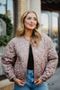 A woman with wavy blonde hair stands outdoors in front of a windowed building, wearing the Katydid Brown Leopard Quilted Jacket over a black top and blue jeans.