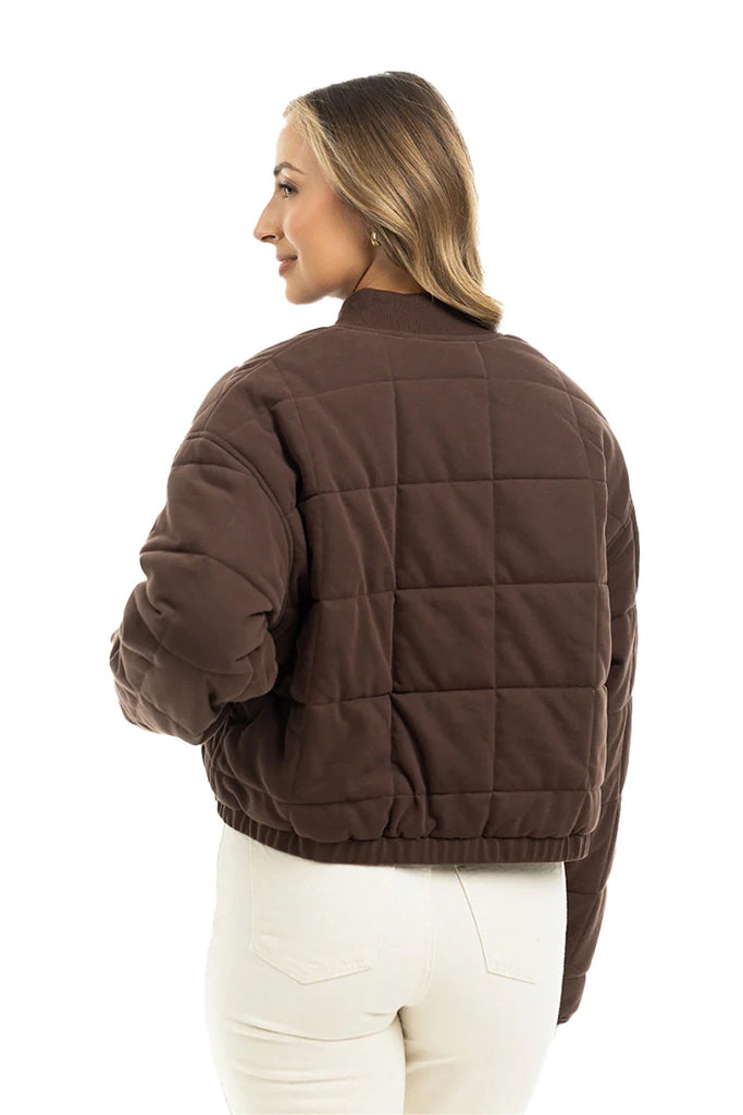 A woman faces away, wearing the Katydid Chocolate Brown Cropped Quilted Bomber Jacket and high-waisted white pants against a white background.