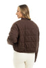 A woman faces away, wearing the Katydid Chocolate Brown Cropped Quilted Bomber Jacket and high-waisted white pants against a white background.