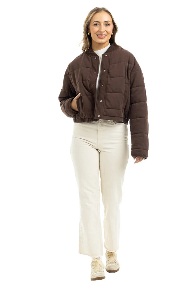A woman smiles wearing the Katydid Chocolate Brown Cropped Quilted Bomber Jacket over a white top, cream pants, and light sneakers, standing against a plain white background.