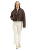 A woman smiles wearing the Katydid Chocolate Brown Cropped Quilted Bomber Jacket over a white top, cream pants, and light sneakers, standing against a plain white background.