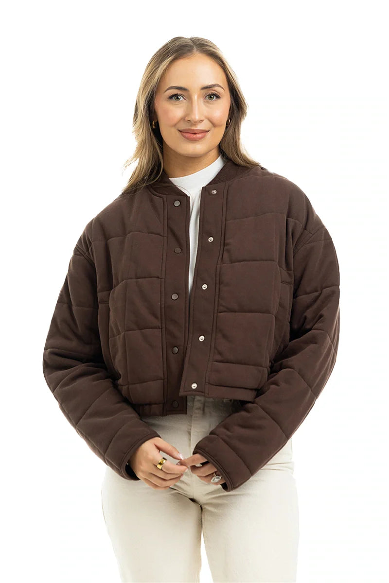 A woman with light brown hair smiles against a white background, wearing the Katydid Chocolate Brown Cropped Quilted Bomber Jacket over a white turtleneck and light pants.