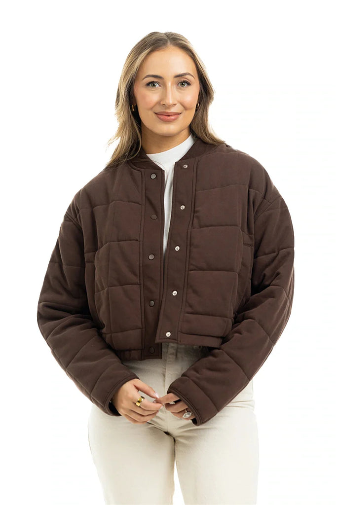 A woman with light brown hair smiles against a white background, wearing the Katydid Chocolate Brown Cropped Quilted Bomber Jacket over a white turtleneck and light pants.