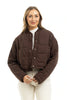 A woman with light brown hair smiles against a white background, wearing the Katydid Chocolate Brown Cropped Quilted Bomber Jacket over a white turtleneck and light pants.