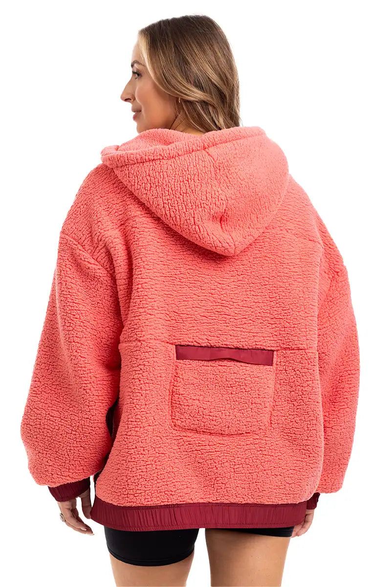 A woman with light brown hair faces away, wearing the Katydid Coral Two-Toned Womens Fleece Pullover with a hood and small back pocket, paired with black shorts.