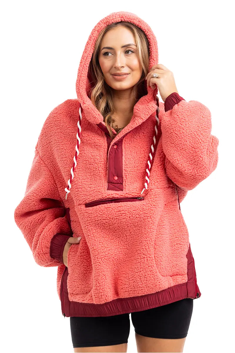 A woman smiles in the Katydid Coral Two-Toned Womens Fleece Pullover, featuring a cozy sherpa design, front zipper, white striped drawstrings, and maroon accents. She pairs it with black shorts and rests one hand on the hoodie.