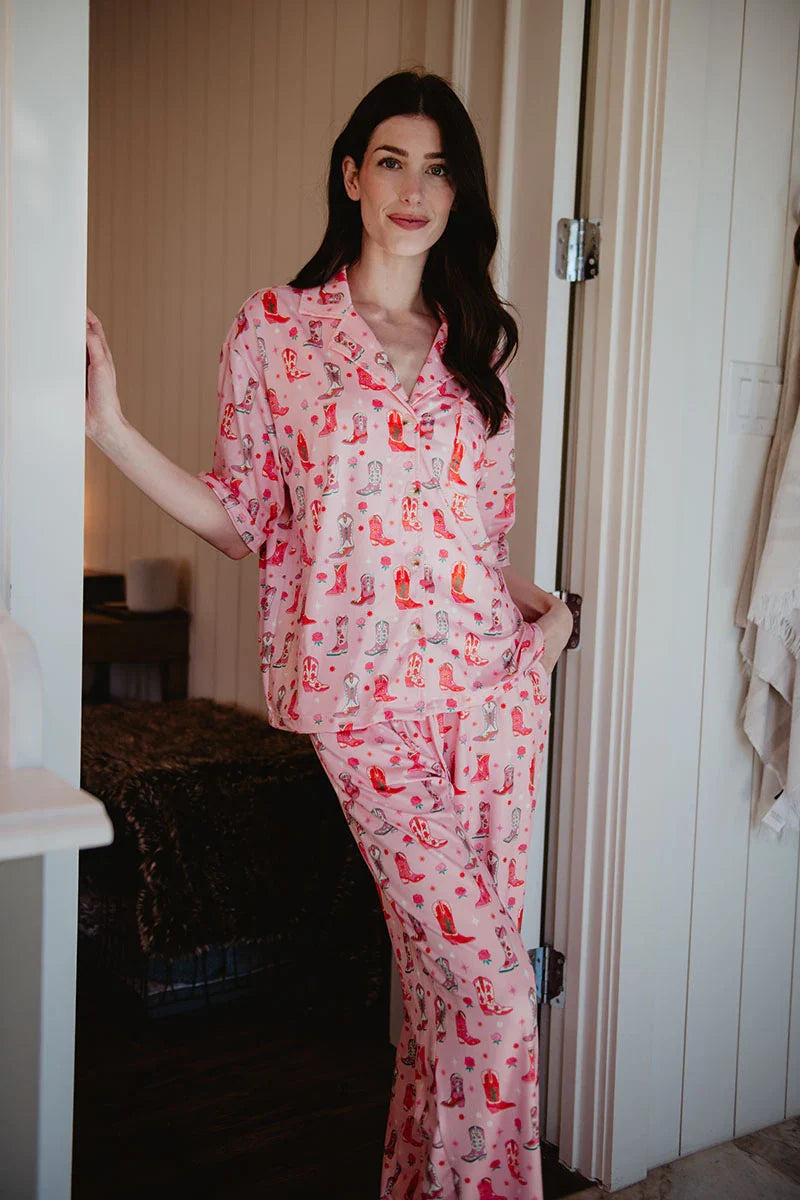 A woman with long dark hair stands in a doorway, smiling in the Katydid Womens Boots  Roses PJ Set—colorful and cozy against white walls.