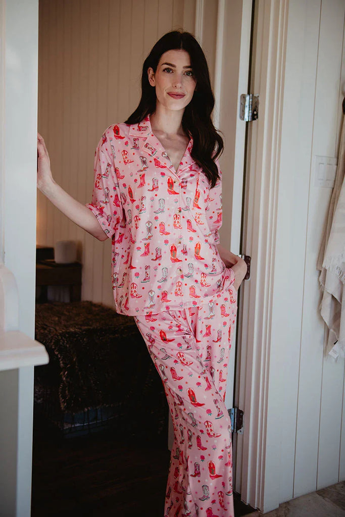 A woman with long dark hair stands in a doorway, smiling in the Katydid Womens Boots  Roses PJ Set—colorful and cozy against white walls.