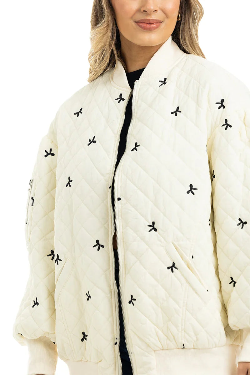 A woman models the Katydid Cream and Black Bows Oversized Quilted Jacket, featuring delicate bow embroidery, against a white background.