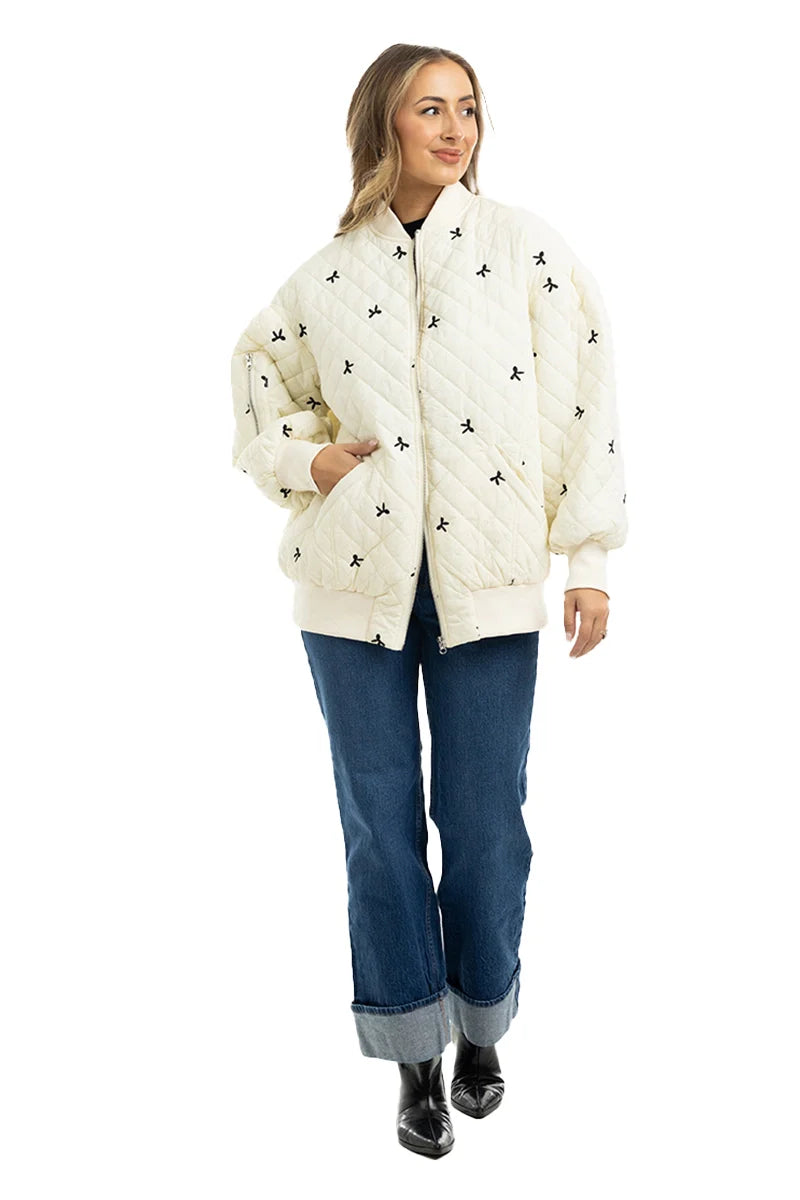 A woman poses against a white background wearing the Katydid Cream and Black Bows Oversized Quilted Jacket, styled with blue cuffed jeans and black ankle boots. She has long, wavy hair and keeps her hands in her jacket pockets.