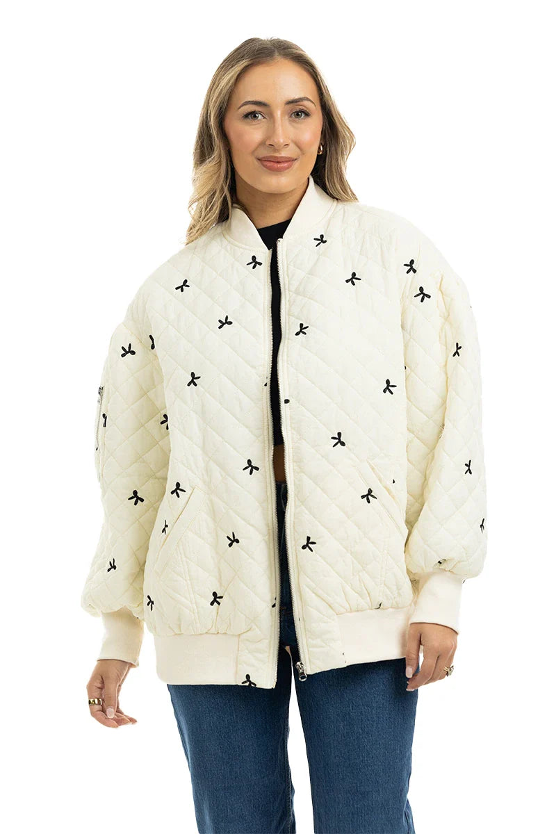 A woman wearing the Katydid Cream and Black Bows Oversized Quilted Jacket stands against a white background, smiling at the camera while pairing it with blue jeans and rings.