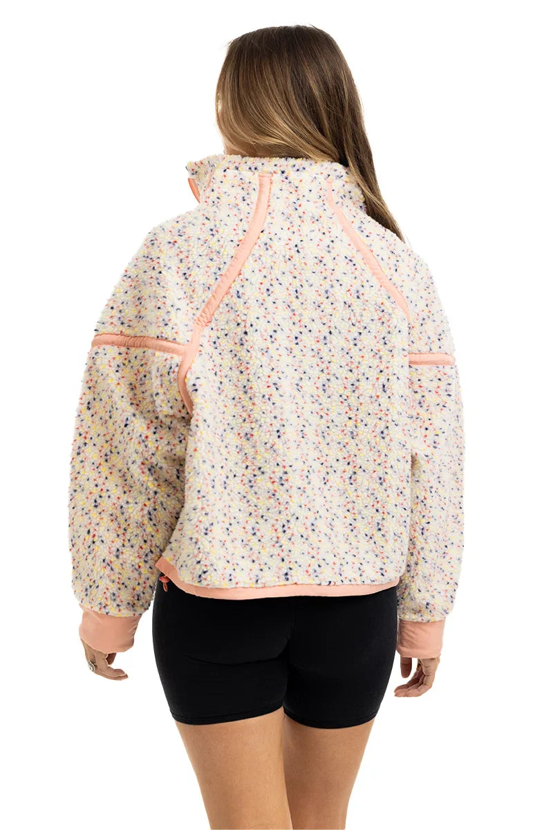 A woman with long brown hair is seen from behind wearing the Katydid Confetti Print Womens Pullover, featuring colorful speckles and light pink trim, paired with black shorts for a cozy, stylish look.