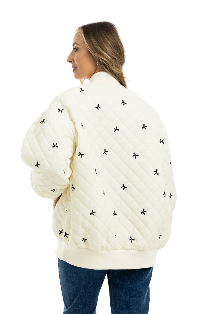 A woman with long, light brown hair models the Katydid Cream and Black Bows Oversized Quilted Jacket, featuring small bow embroidery. She faces away with her head to the side and wears blue jeans.
