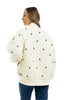 A woman with long, light brown hair models the Katydid Cream and Black Bows Oversized Quilted Jacket, featuring small bow embroidery. She faces away with her head to the side and wears blue jeans.