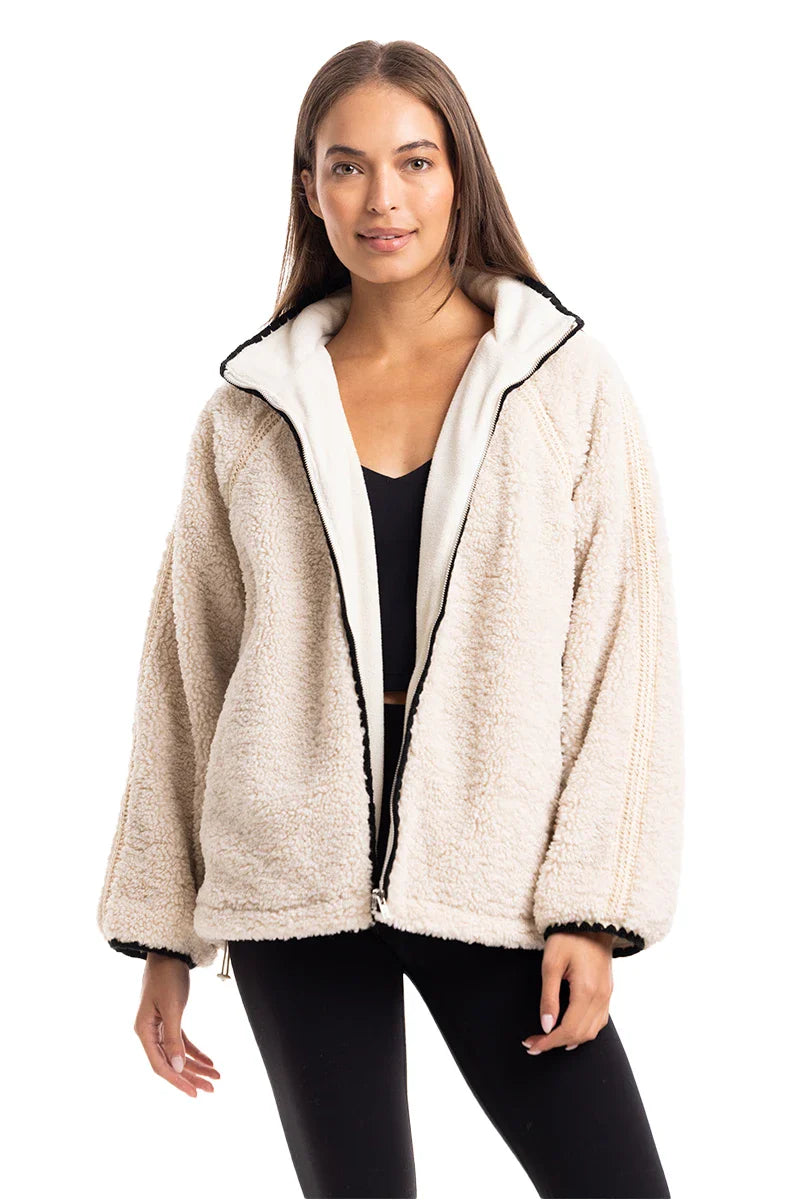 A woman with long brown hair models the Katydid Cream Womens Sherpa Lace Trim Jacket, styled oversized over a black crop top and leggings, standing against a plain white background.