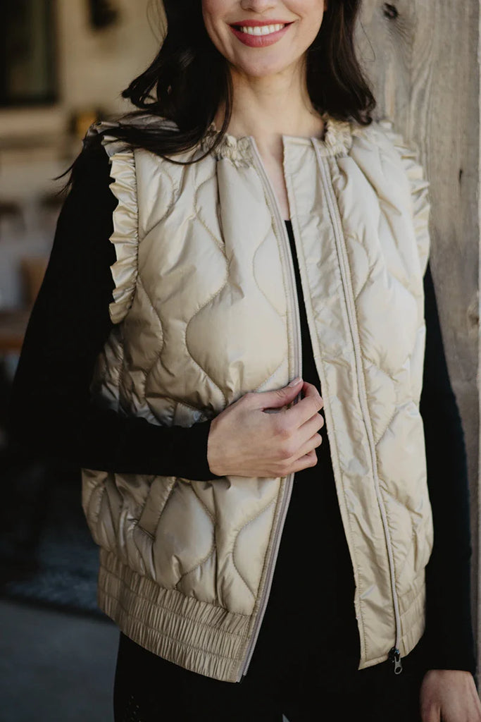 A woman stands indoors, smiling and touching the ruffled shoulder of her Katydid Soft Tan Metallic Quilted Vest, styled over a black long-sleeve top.
