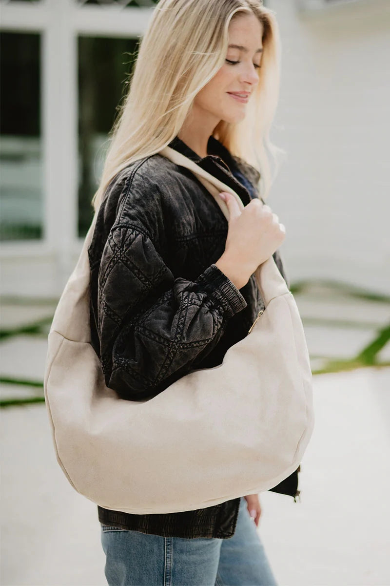 A woman with long blonde hair, in a dark denim jacket and blue jeans, stands outdoors smiling as she carries the Katydid Cream Versatile Suede Tote Bag over her shoulder.