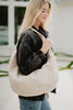 A woman with long blonde hair, in a dark denim jacket and blue jeans, stands outdoors smiling as she carries the Katydid Cream Versatile Suede Tote Bag over her shoulder.
