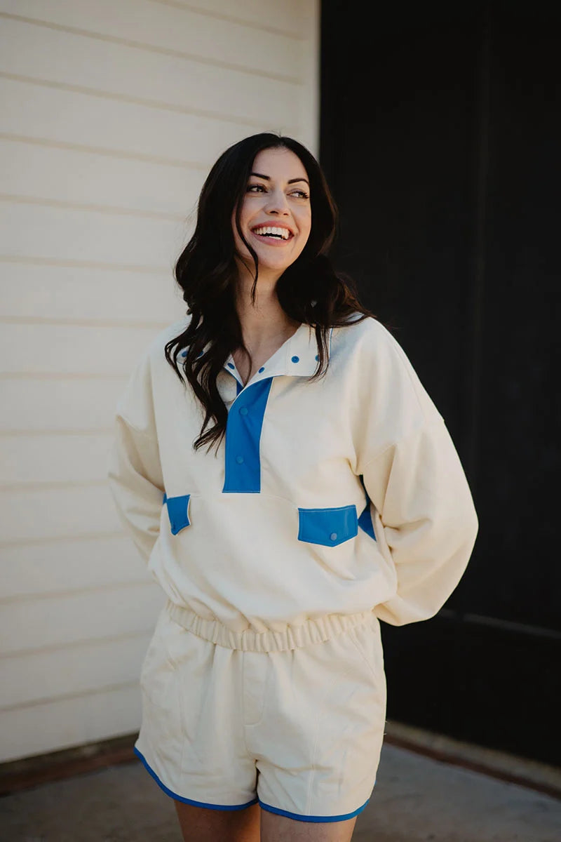 A woman with long dark hair smiles outdoors in a Katydid Yellow  Blue Quarter Snap Athletic Set—cream-colored, accented with blue. She wears a matching long-sleeve top and shorts, standing before a white wall and dark doorway.