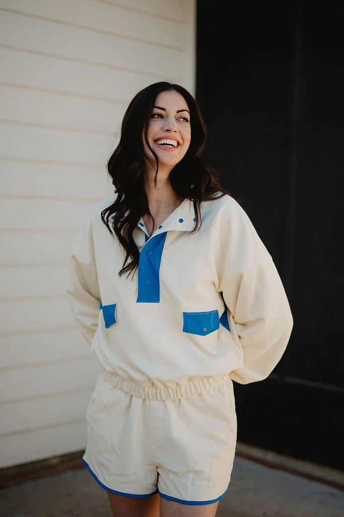 A woman with long dark hair smiles outdoors in a Katydid Yellow  Blue Quarter Snap Athletic Set—cream-colored, accented with blue. She wears a matching long-sleeve top and shorts, standing before a white wall and dark doorway.