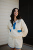 A woman with long dark hair smiles outdoors in a Katydid Yellow  Blue Quarter Snap Athletic Set—cream-colored, accented with blue. She wears a matching long-sleeve top and shorts, standing before a white wall and dark doorway.
