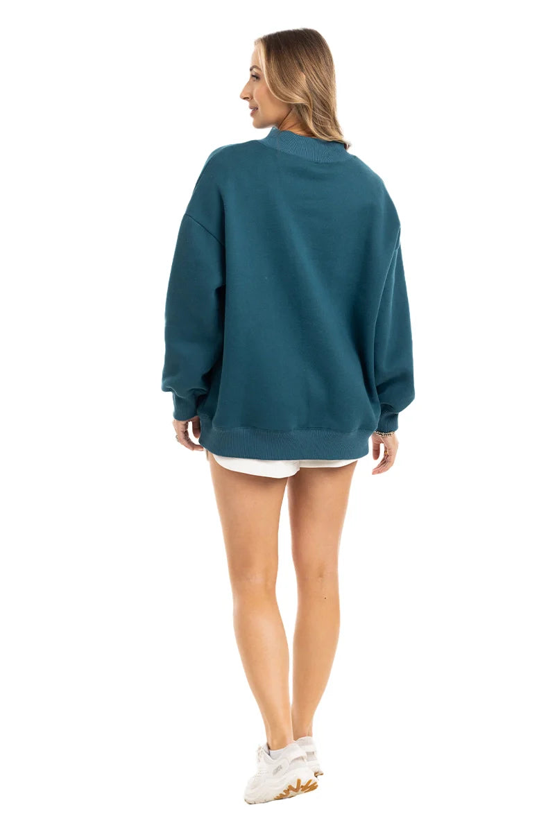 A woman with light brown hair smiles, facing away, in the Katydid Deep Teal Essential Mockneck Sweatshirt. She pairs it with white shorts and sneakers for a perfect fall look against a plain white background, her left hand slightly extended.