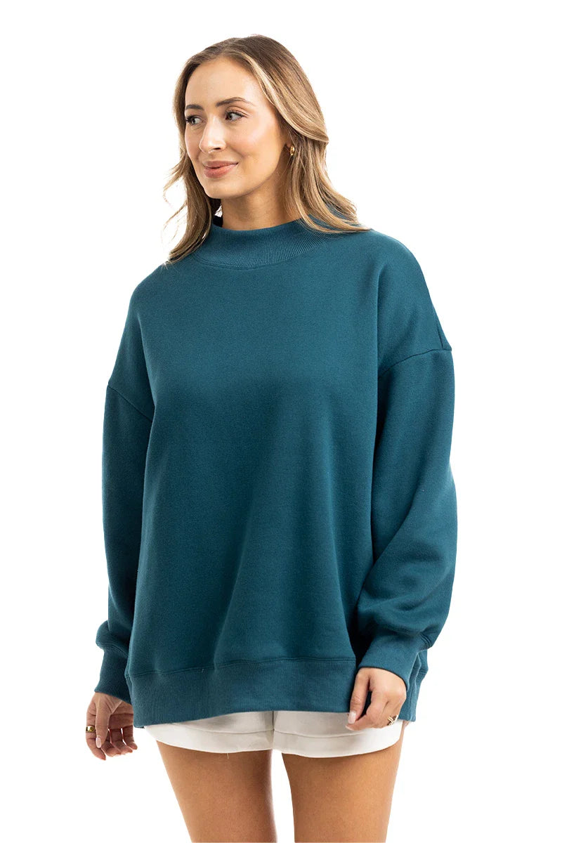 A woman with shoulder-length light brown hair smiles softly, modeling the Katydid Deep Teal Essential Mockneck Sweatshirt against a white background. She pairs the oversized top with white shorts for a cozy fall look.