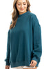 A woman with long, light brown hair smiles gently in a Katydid Deep Teal Essential Mockneck Sweatshirt and light pants against a white background—a cozy, stylish pick for fall.