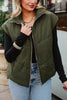 A woman stands outdoors in a Katydid Olive Corduroy Puffer Vest over a black long-sleeve top, light blue jeans, gold jewelry, and neutral nail polish. Her face is partially visible.