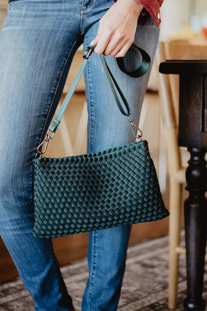 A person in blue jeans holds the Katydid Forest Green Woven Everywhere Crossbody with shoulder strap, standing indoors near a wooden table and chair.