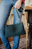 A person in blue jeans holds the Katydid Forest Green Woven Everywhere Crossbody with shoulder strap, standing indoors near a wooden table and chair.