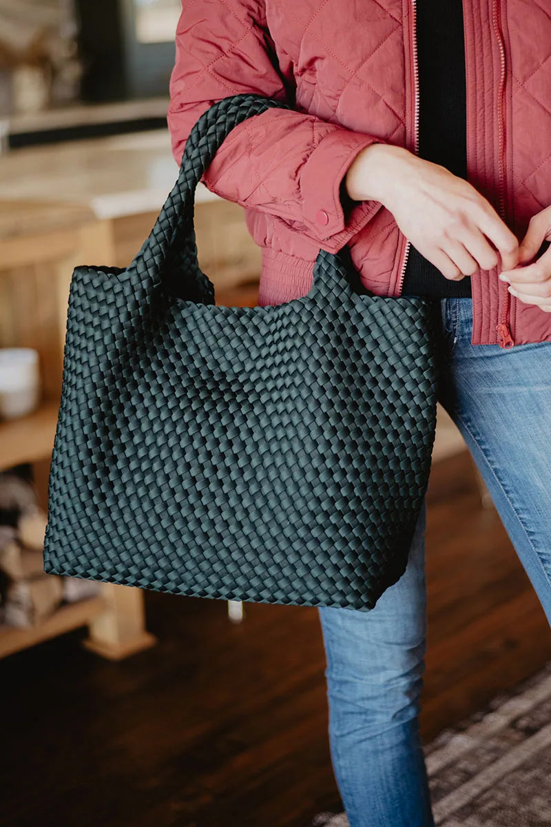 A person in a red jacket and jeans holds the Katydid Forest Green Woven Tote Bag by its handles indoors.