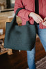 A person in a red jacket and jeans holds the Katydid Forest Green Woven Tote Bag by its handles indoors.