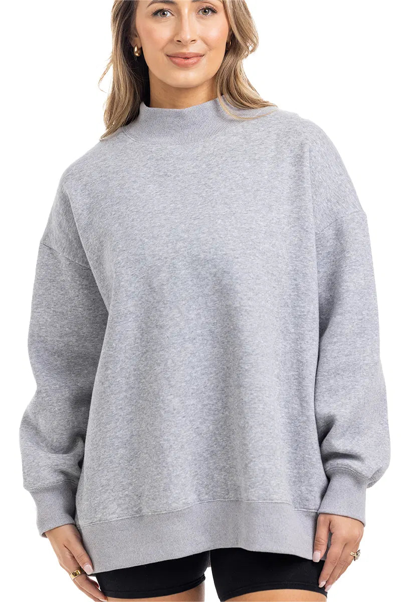 A woman wears the Katydid Heather Essential Mockneck Sweatshirt, a loose-fitting, heather gray pullover with long sleeves—an ideal cozy piece for your fall wardrobe.