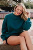 A smiling woman with long blonde hair sits outdoors by a fountain, wearing the Katydid Deep Teal Essential Mockneck Sweatshirt and black shorts as a stylish fall outfit, with one hand touching her hair.